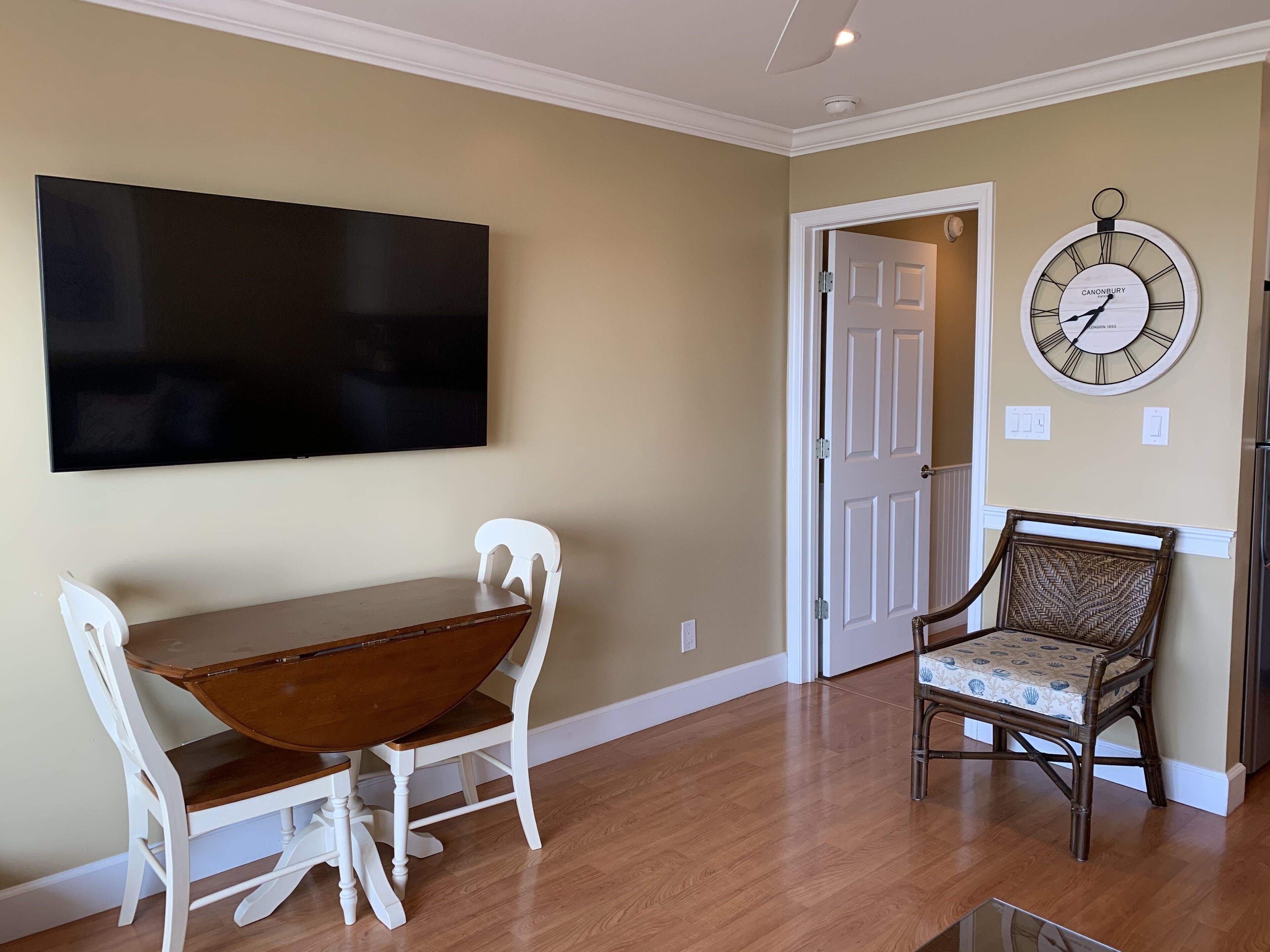 A dining area with a wooden table and chairs, a flat-screen TV mounted on the wall, and a wall clock.