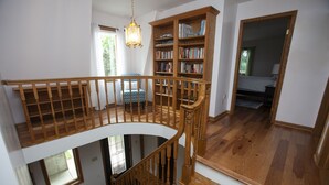 A spacious room with a wooden staircase, a large bookshelf, and a view of a bedroom through an open door. - Peaceful Retreat for Families/Couples/Groups - All Sports Lake w/ Kayaks & SUP's (Cadillac)