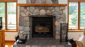 A stone fireplace with a wooden mantel, a black TV stand, and a leather chair. - Peaceful Retreat for Families/Couples/Groups - All Sports Lake w/ Kayaks & SUP's (Cadillac)