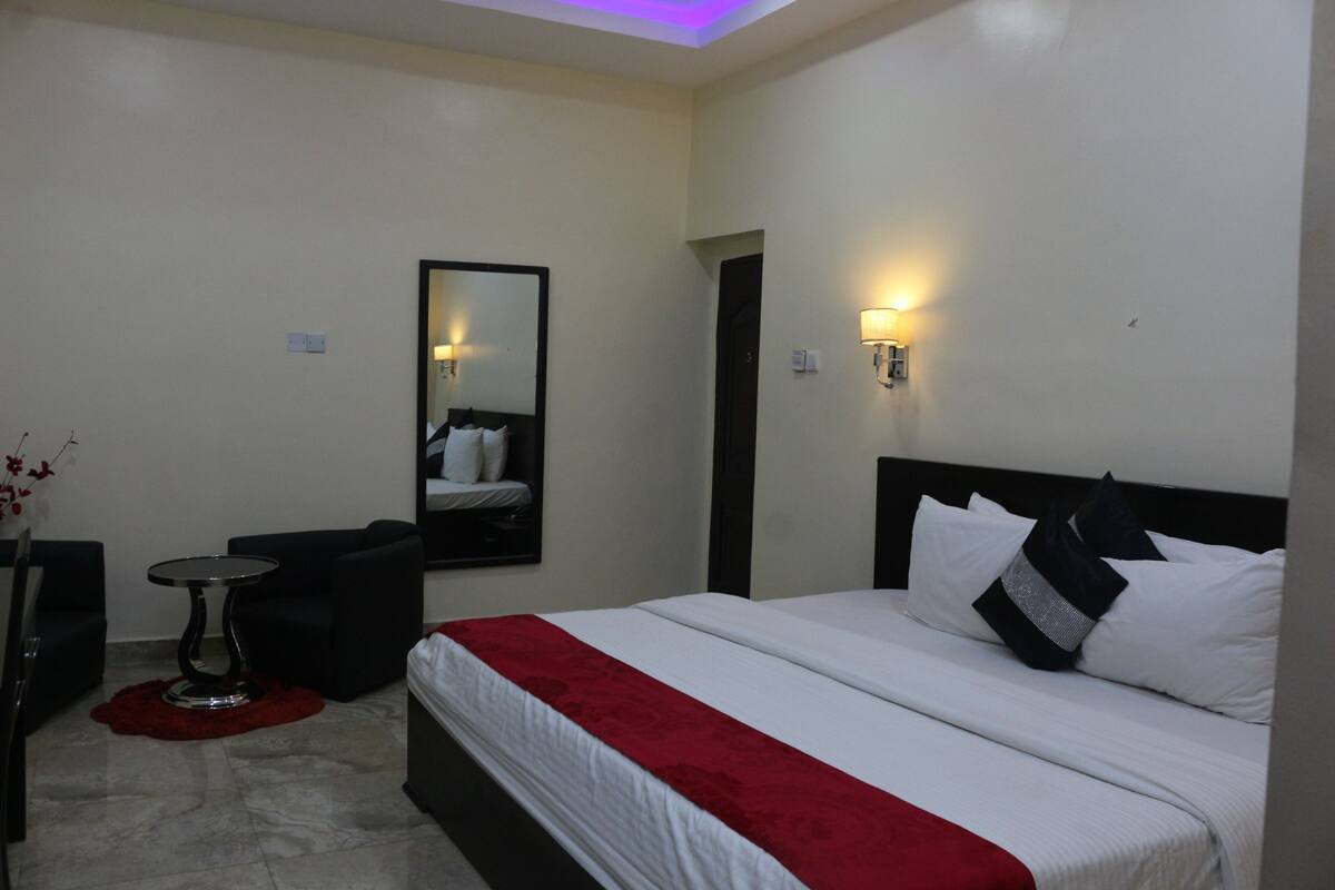 A hotel room with a bed, a small table, a chair, a mirror, and two wall-mounted lights.