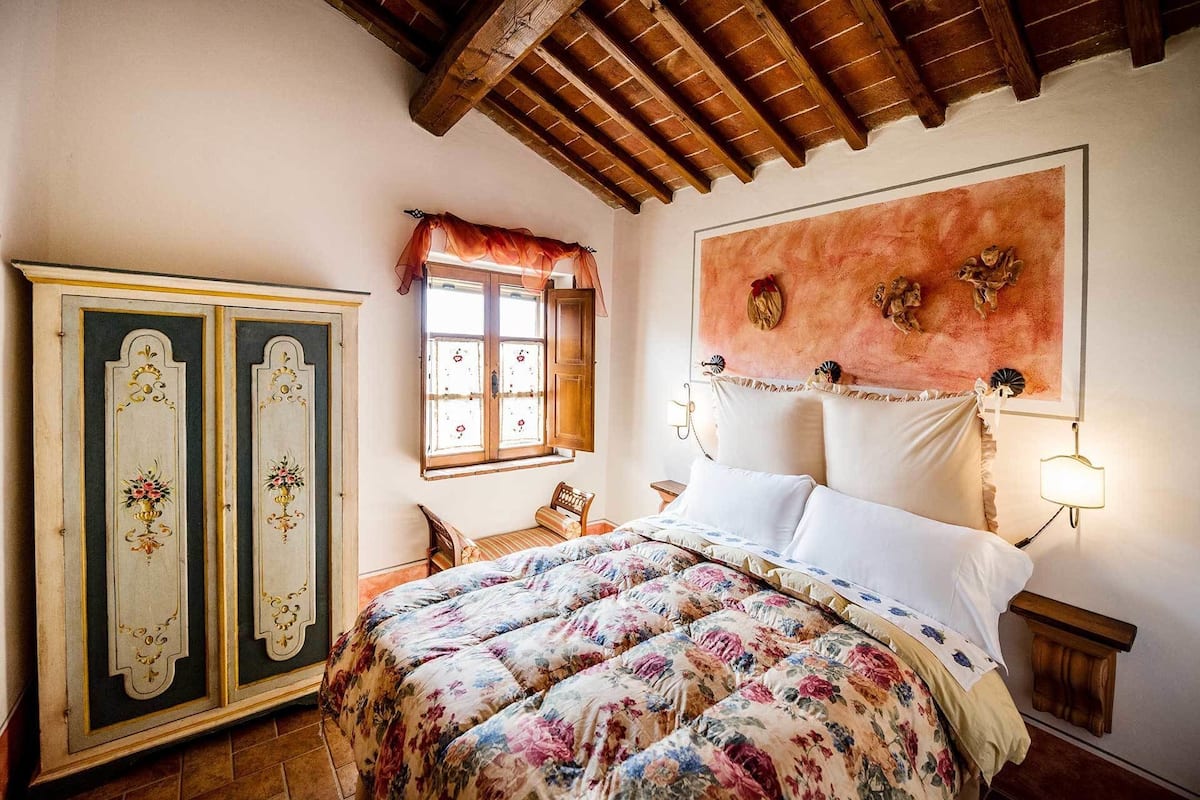 Ein Schlafzimmer mit Holzboden, einem Bett mit floralem Bettbezug, einem Nachttisch, einem Stuhl und einem Kleiderschrank mit dekorativen Paneelen.