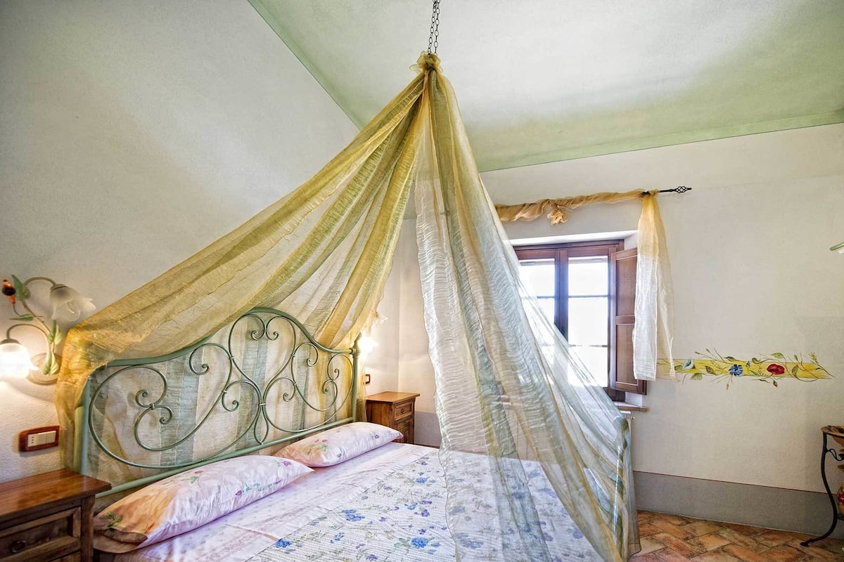 Ein Schlafzimmer mit einem Himmelbett, einem Holfnachtkästchen, einem Nachttisch und einem Fenster mit Vorhängen.