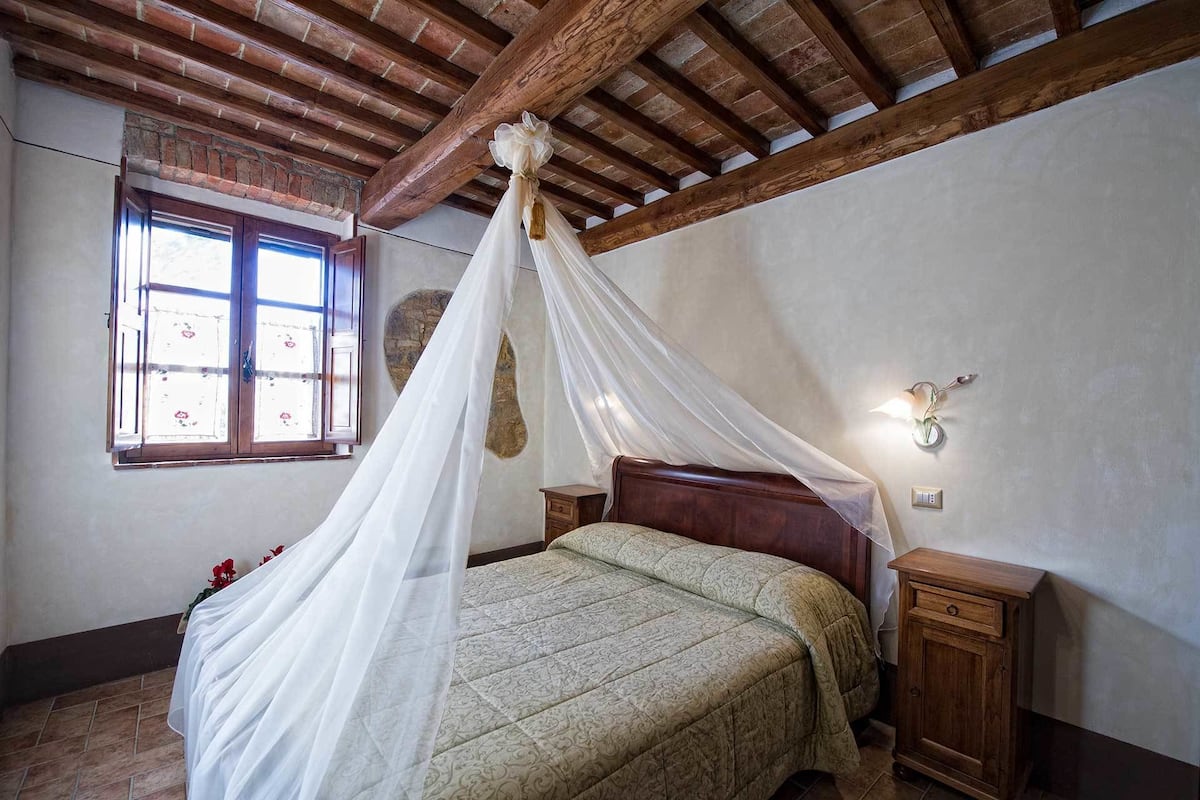 Ein Schlafzimmer mit Holzbalkendecke, einem Himmelbett, einem Nachttisch, einer Wandleuchte und einem Fenster mit Aussicht.