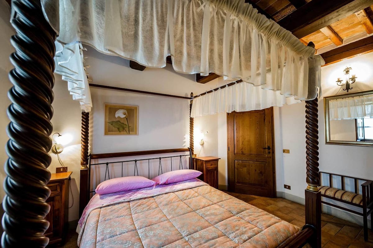 Ein Schlafzimmer mit einem Himmelbett, einem Holz-Kopfstück und einem Baldachin. Es gibt einen Schrank aus Holz und einen Spiegel an der Wand.