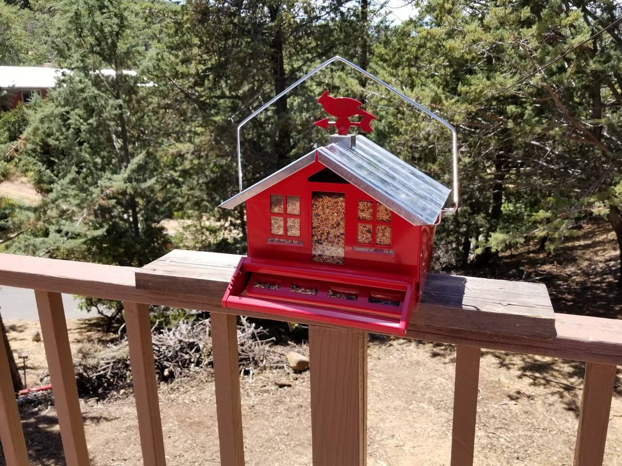 A red birdhouse on a wooden railing with a metal roof, surrounded by trees.