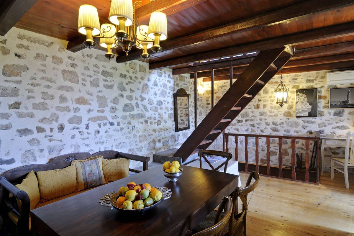 A rustic dining area with a stone wall, wooden ceiling beams, and a staircase leading to an upper level. A wooden table with a bowl of fruit is set for dining.