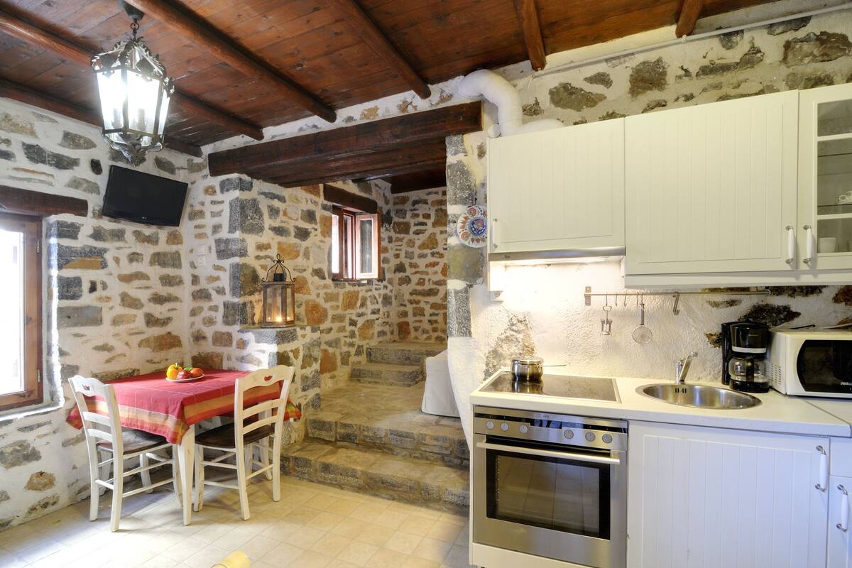 A rustic kitchen with stone walls, wooden beams, a dining table with chairs, a stove, a sink, and a microwave.