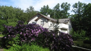 A two-story house with a sloped roof, surrounded by lush greenery and purple flowering bushes.