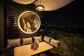 Un área de estar moderna al aire libre con una lámpara circular, una mesa con una botella y vasos, y vistas de la ciudad iluminada por la noche.