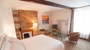 A hotel room with a brick wall, a bed with white linens, a chair, a table, and a radiator. - Le Relais du Beffroi (Saint-Riquier)