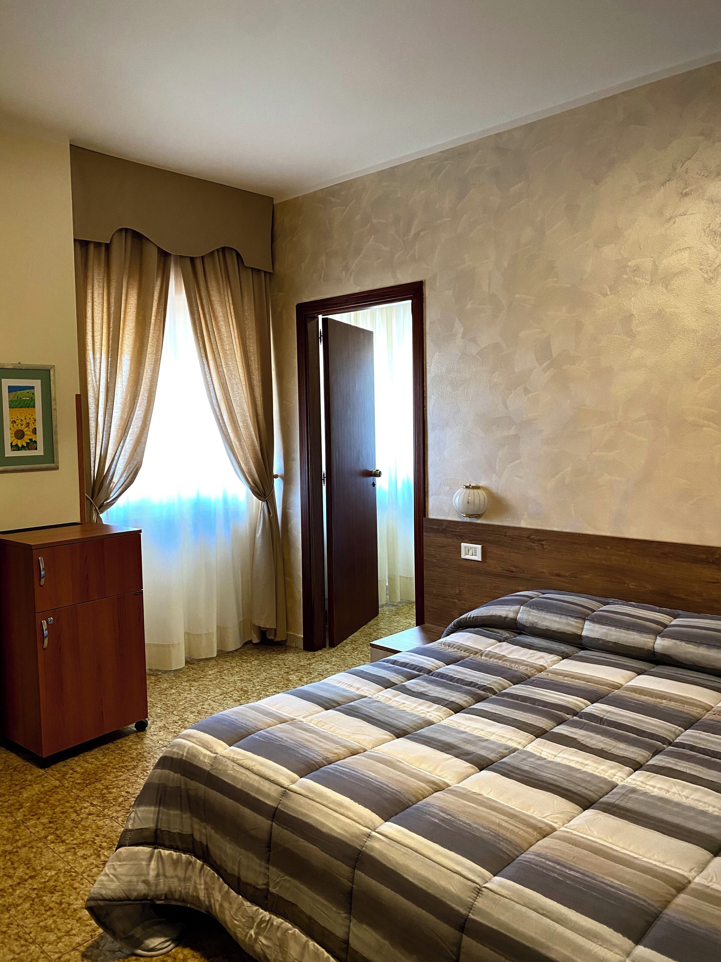 A hotel room with a bed, a wooden wardrobe, a door, and a window with curtains.