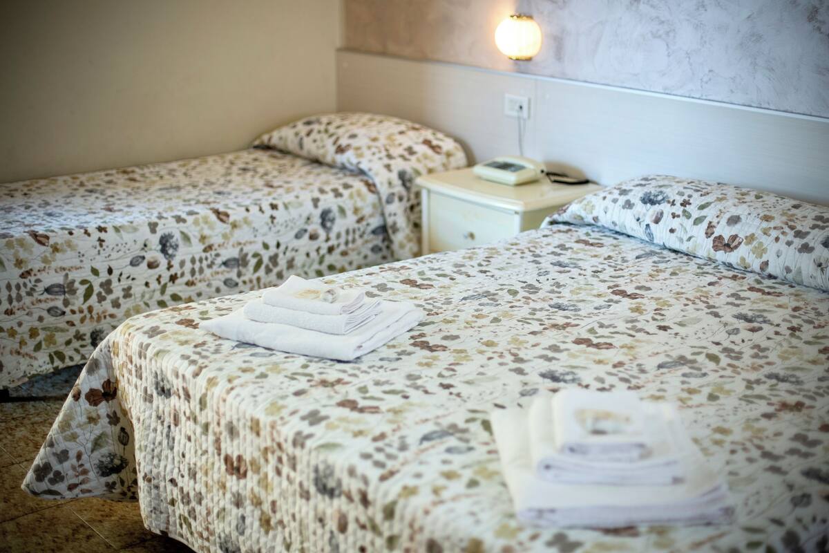 A hotel room with two beds, each with a floral-patterned bedspread and pillows. There is a bedside table with a lamp and a telephone.