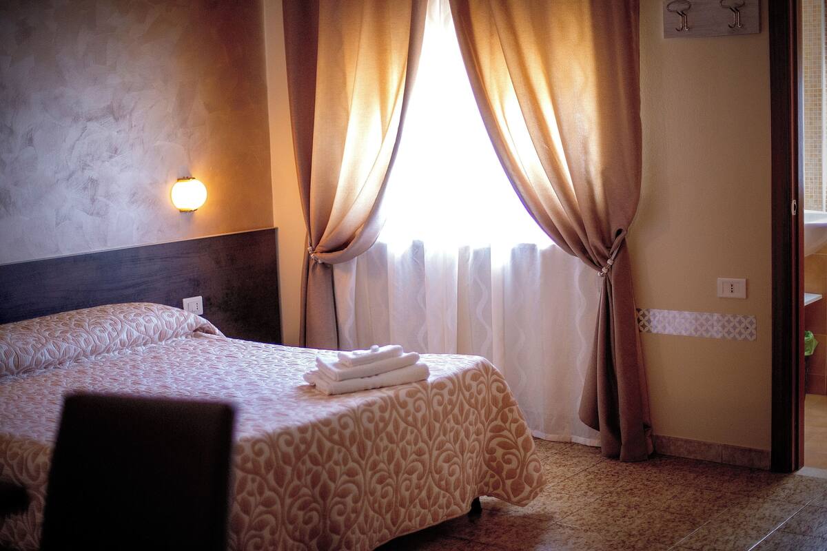 A hotel room with a bed, curtains, a wall-mounted light, and a door.