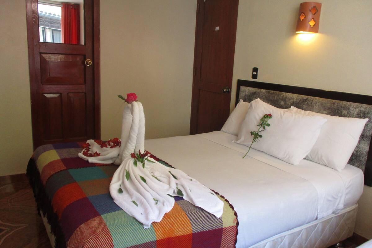 Habitación de hotel con una cama, una manta colorida, almohadas blancas y una lámpara fijada a la pared.