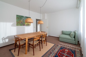 A dining area with a wooden table and chairs, a woven pendant light, a colorful rug, and a green sofa.