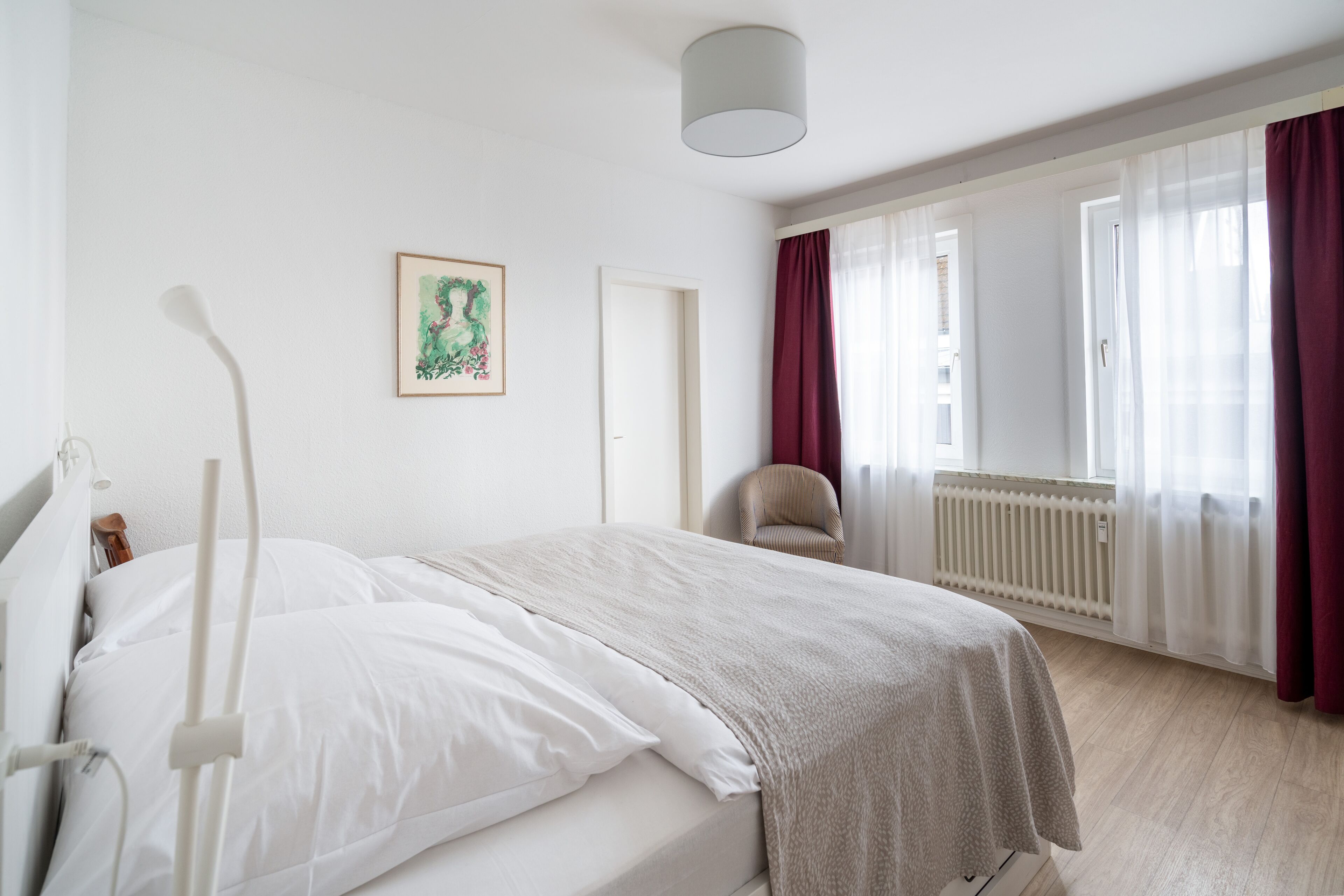 A bedroom with a bed, a chair, a radiator, and a window with curtains.