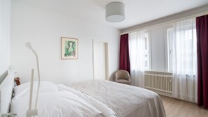 Un dormitorio con cama, una silla, un radiador y una ventana con cortinas.