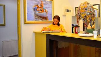 A reception desk with a person writing, a framed painting of a boat scene, and a decorative glass sculpture.