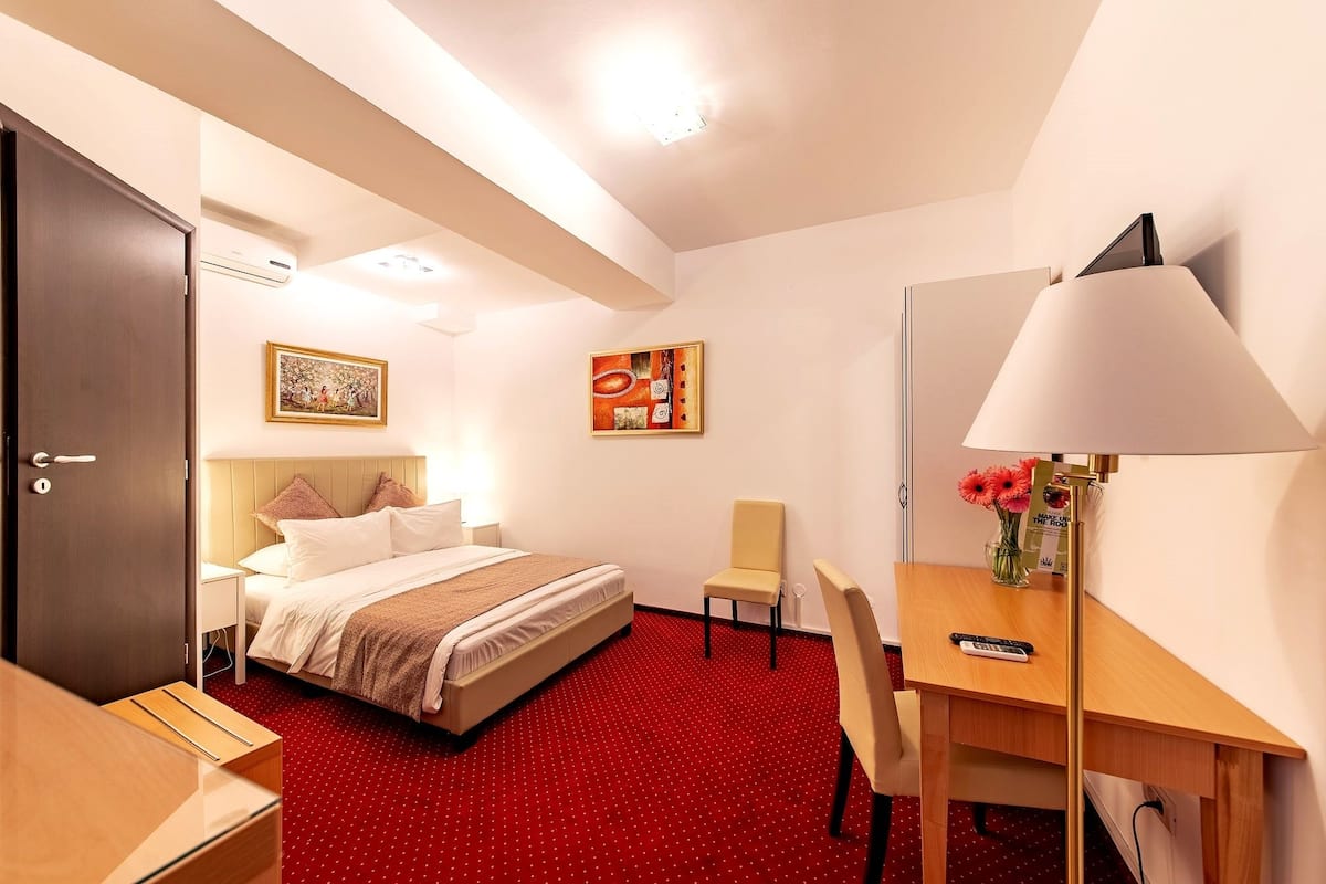 A hotel room with a bed, a desk, a chair, a lamp, and a vase with flowers.
