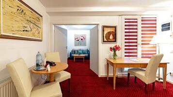 Hotelkamer met rood tapijt, een eettafel met stoelen, een bank, een schilderij en een raam met gestreepte jaloezieën.