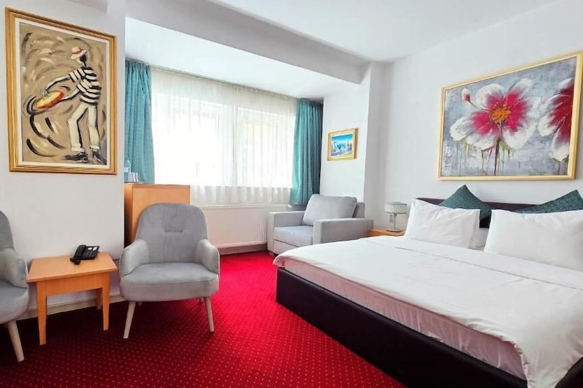 A hotel room with a bed, two armchairs, a small table with a phone, a radiator, and a window with curtains.