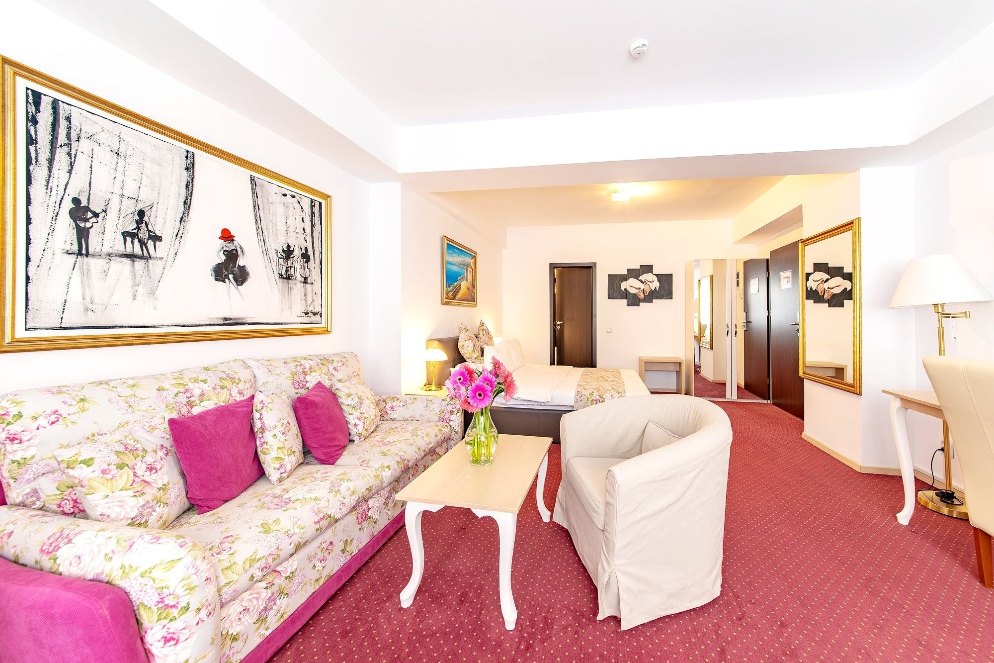 Une chambre d’hôtel avec un canapé à motifs floraux, un fauteuil blanc, une petite table sur laquelle est posé un vase de fleurs, un tableau au mur et une porte qui donne sur une autre pièce.