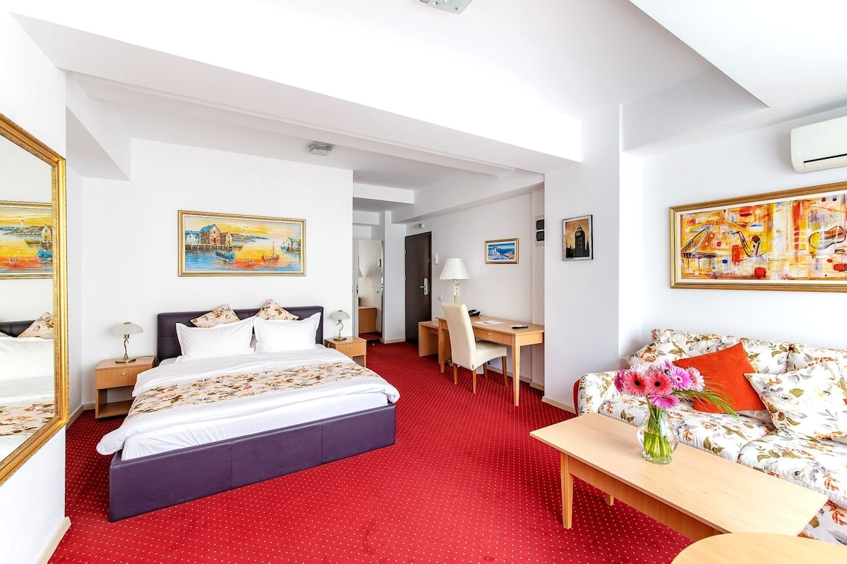 A hotel room with a bed, desk, and sofa. There are paintings on the walls and a red carpet.