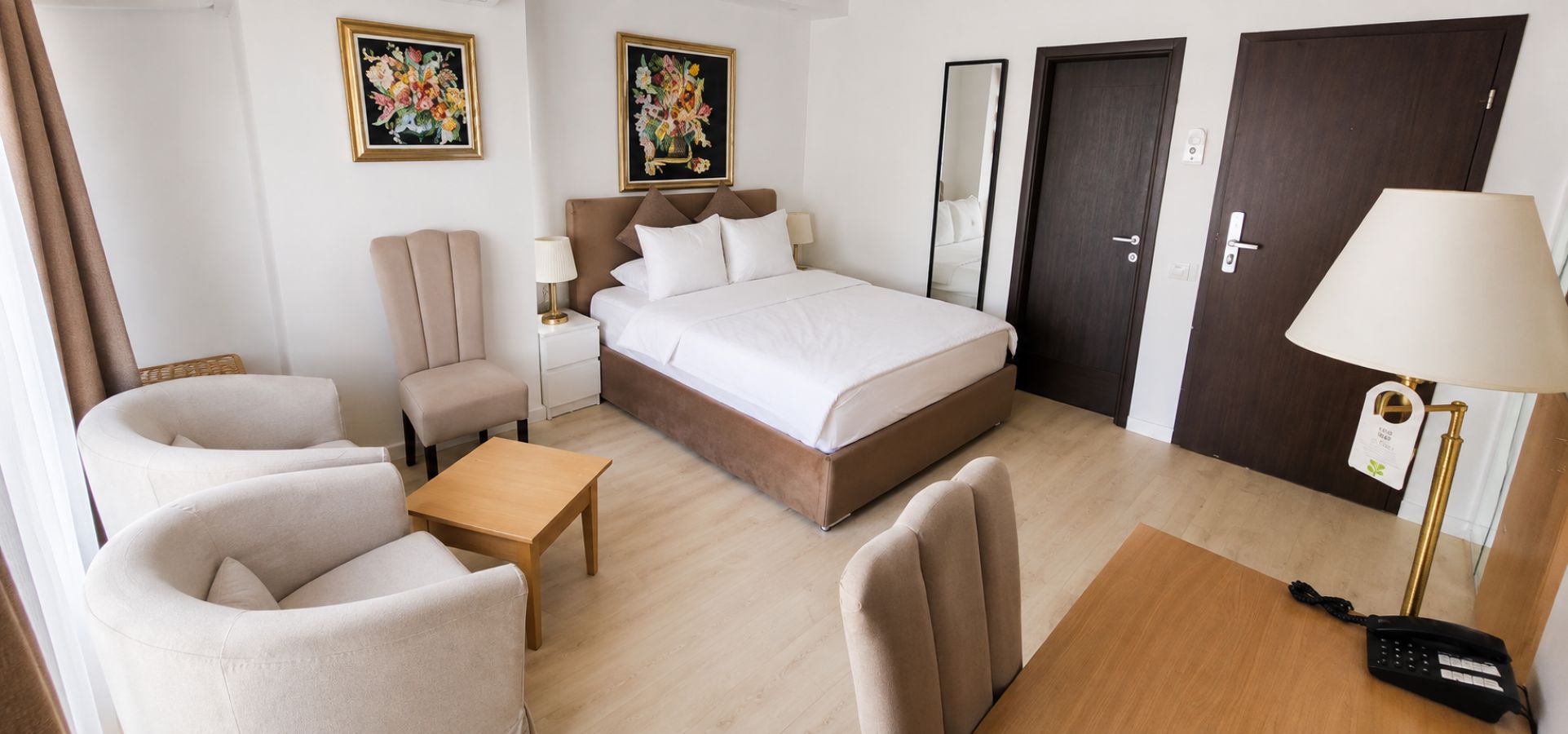 A hotel room with a bed, two armchairs, a small table, a bedside table with a lamp, a mirror, and two doors.