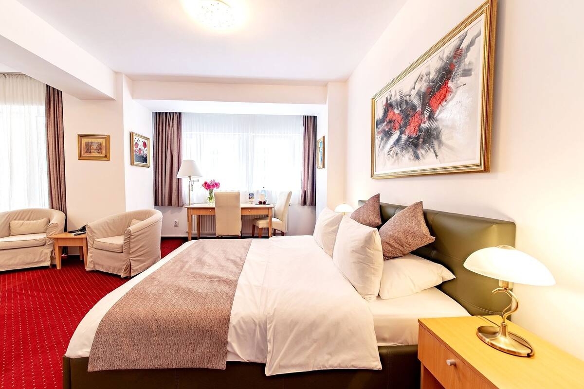 A hotel room with a bed, a desk, a chair, a lamp, and artwork on the wall.