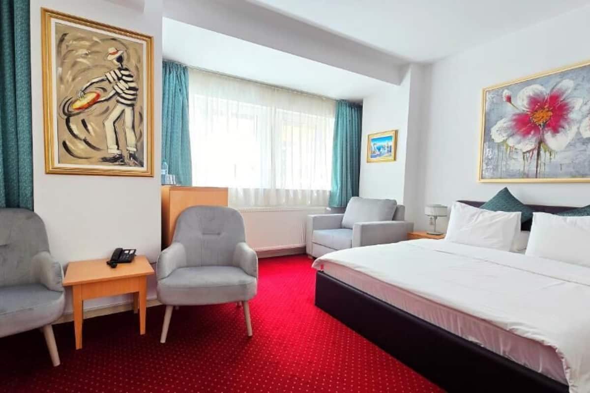 A hotel room with a bed, two armchairs, a small table with a phone, a painting on the wall, and a window with curtains.
