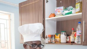 Un chef con uniforme blanco de pie en una cocina con armarios de madera, encimera de granito y varios electrodomésticos.