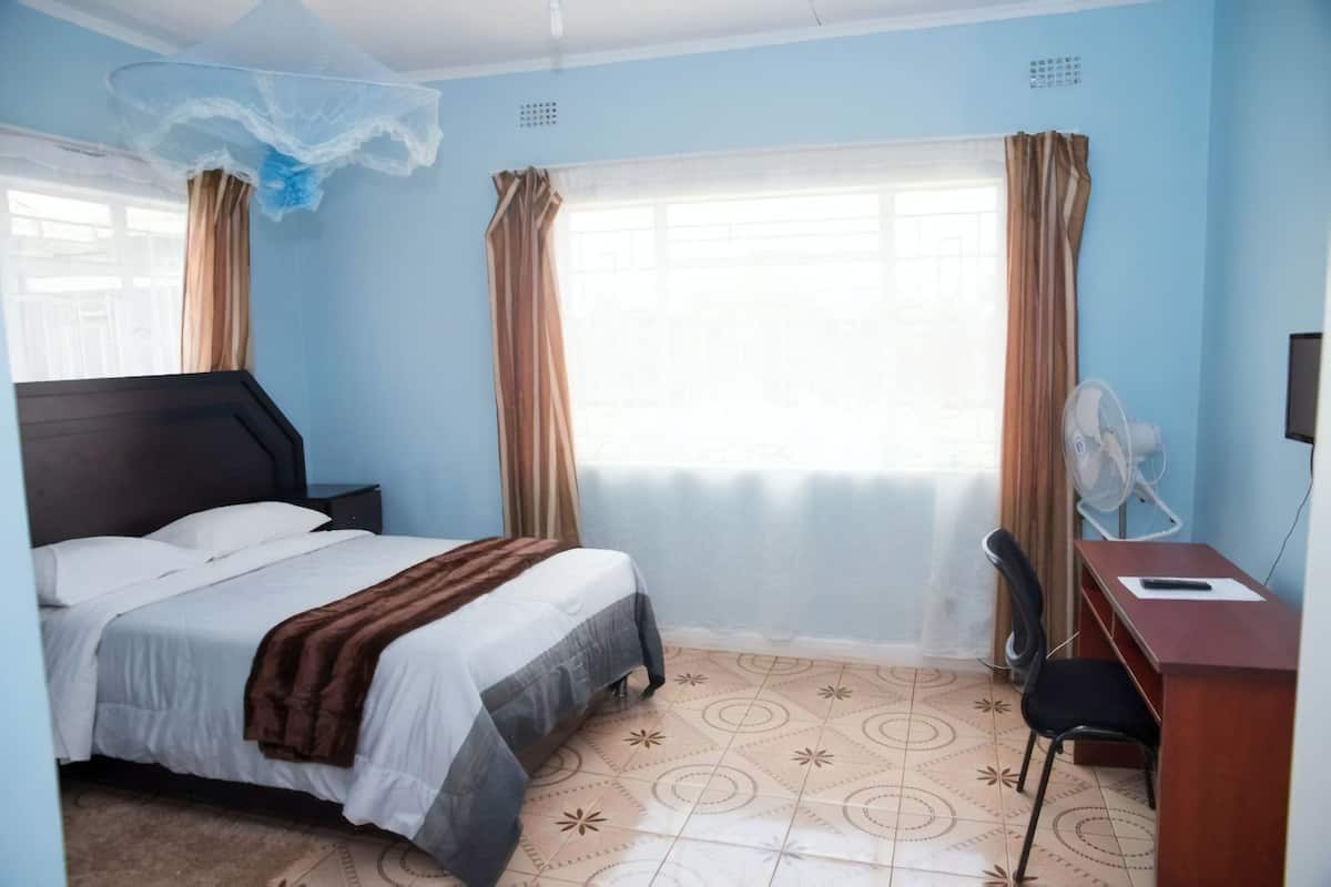 Quarto de hotel com cama, escrivaninha, cadeira, ventilador e janela com cortinas.
