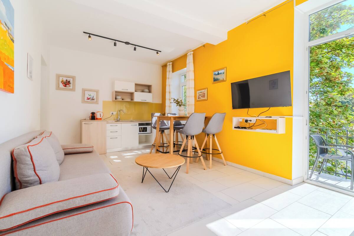 A modern living space with a sofa, dining table, chairs, a TV, and a kitchen area.