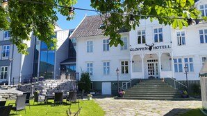 Ein Hotel mit weißer Fassade, einer Steintreppe zum Eingang, Außenbestuhlung mit Stühlen und Tischen und einem klaren Himmel.
