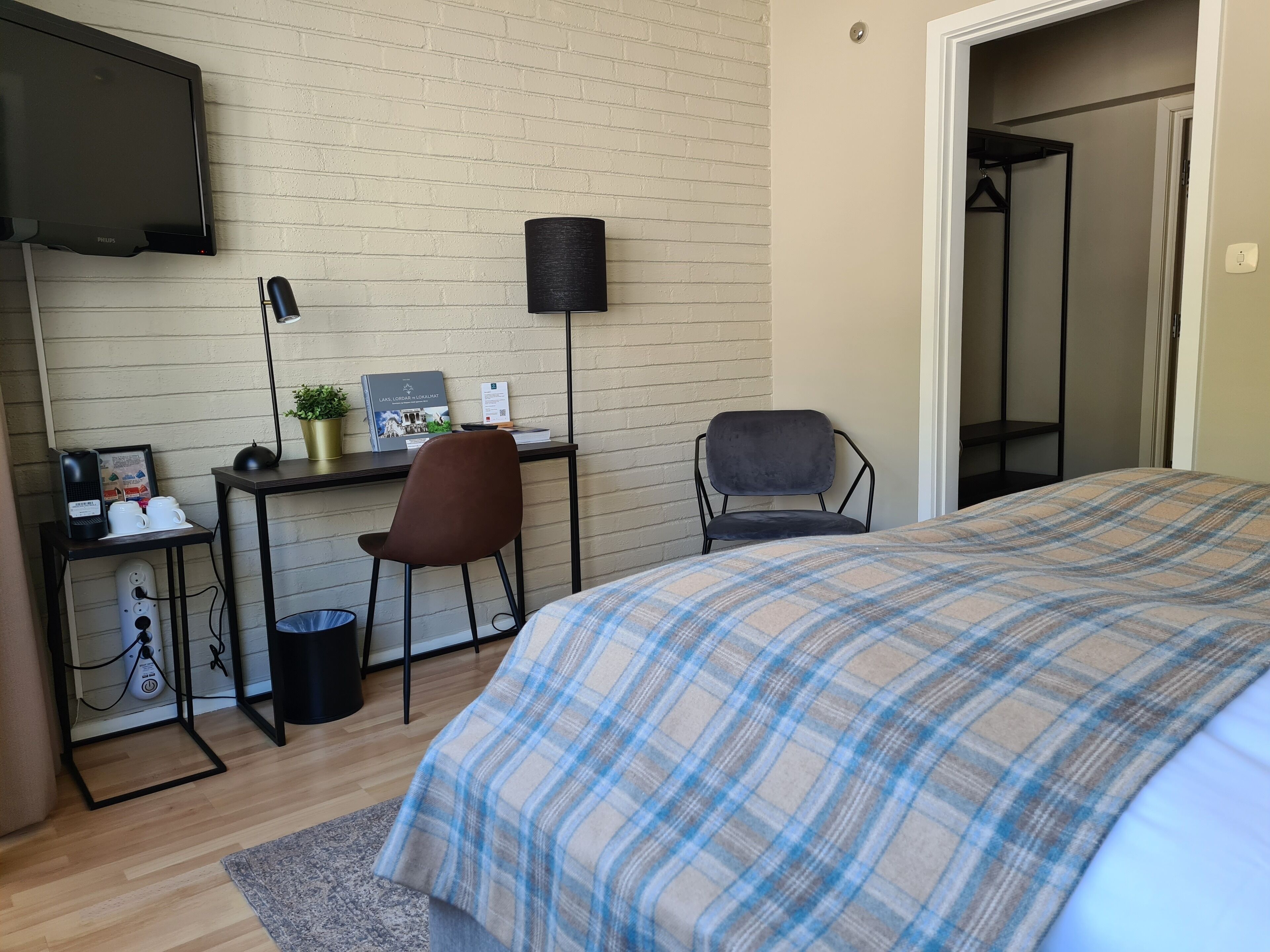 A bedroom with a bed, a desk with a chair, a television, and a small table with a plant and a framed picture.