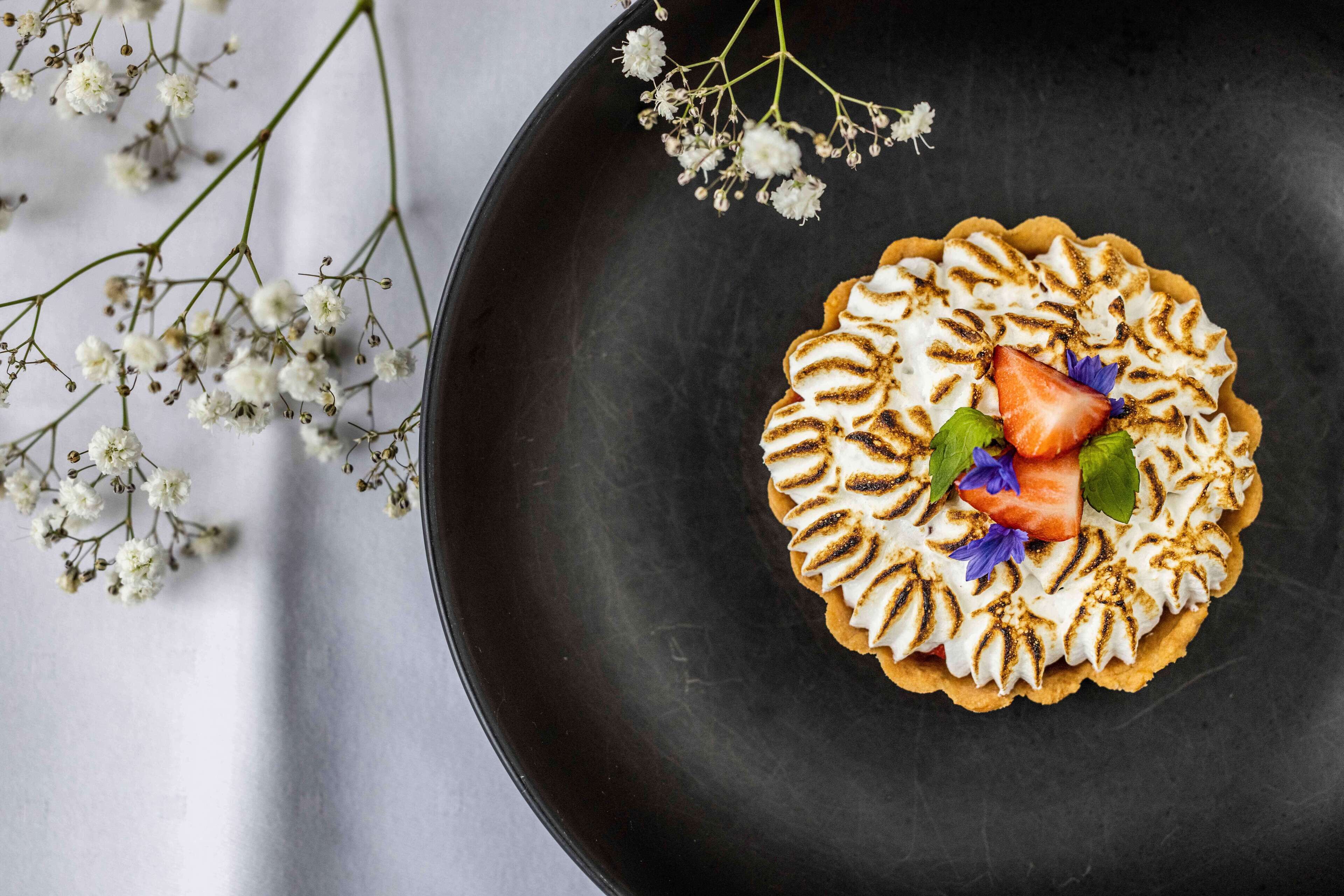 A dessert on a dark plate with a floral arrangement beside it.