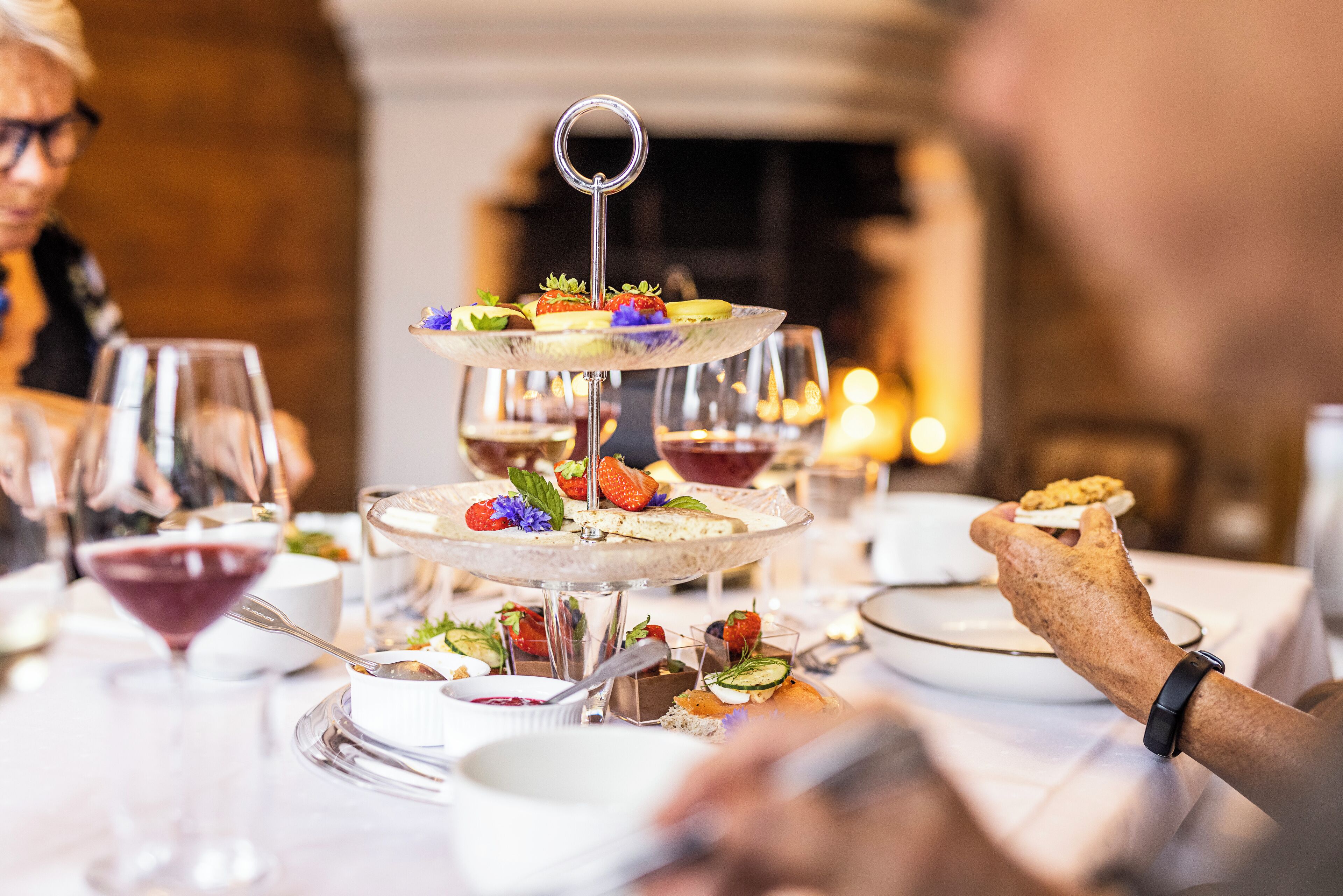 Un présentoir à plusieurs niveaux, garni de diverses pâtisseries et de fruits, entouré de verres à vin et de couverts sur une table.