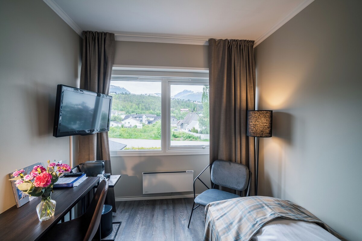 Ein Hotelzimmer mit Bett, Schreibtisch mit Computer, Stuhl, Fernseher und einem Fenster mit Vorhängen.