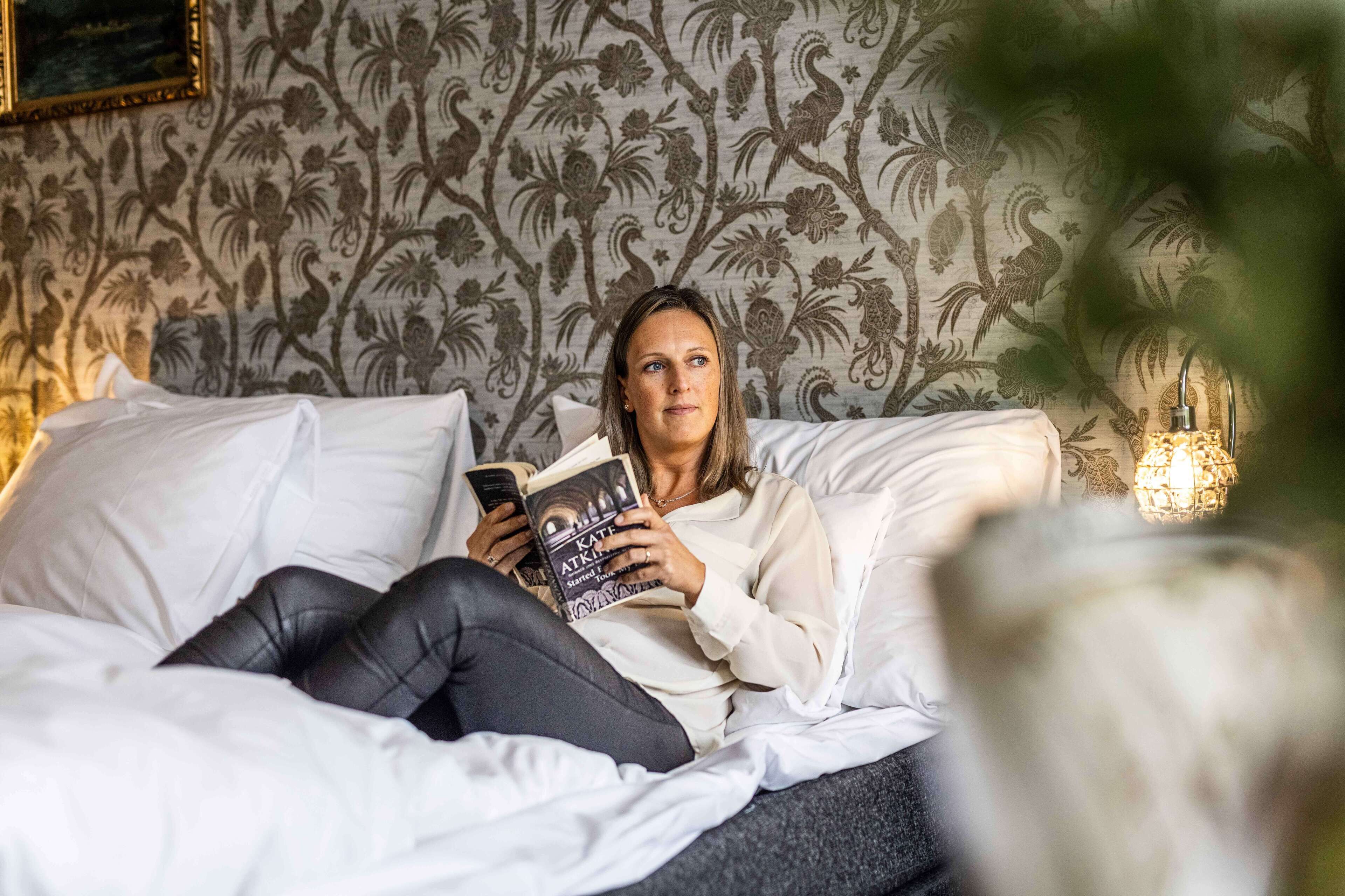 Uma pessoa deitada na cama, lendo um livro, com papel de parede estampado.