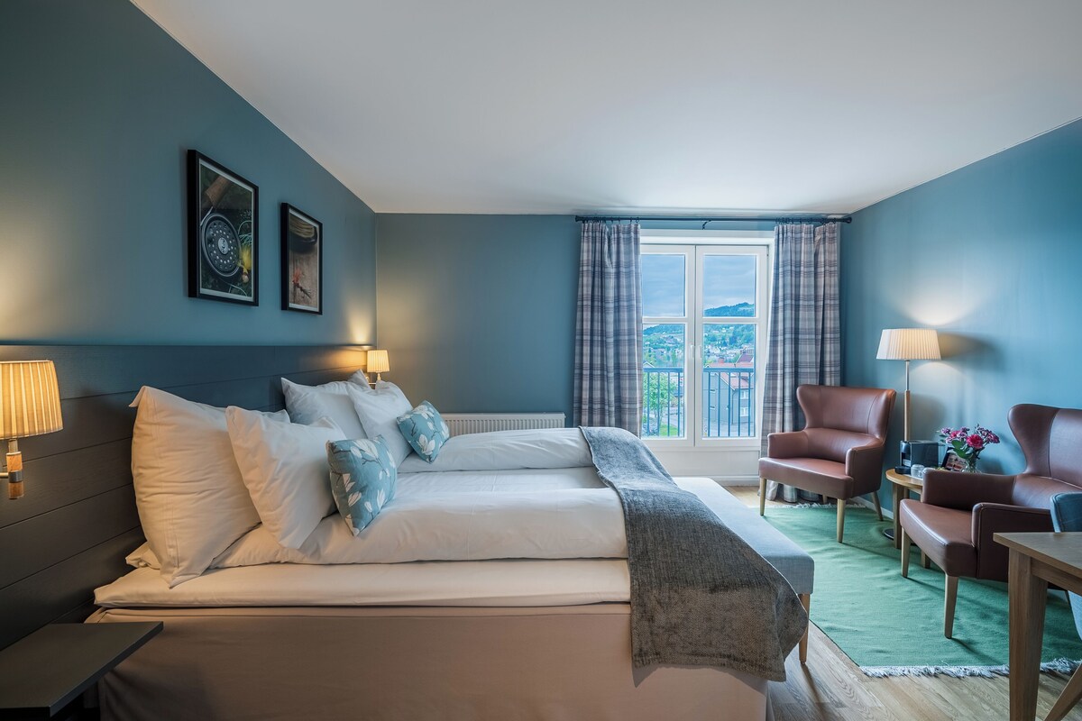 Ein Schlafzimmer mit Bett, zwei Stühlen, einem Tisch, einem Fenster mit Vorhängen und Bildern an der Wand.