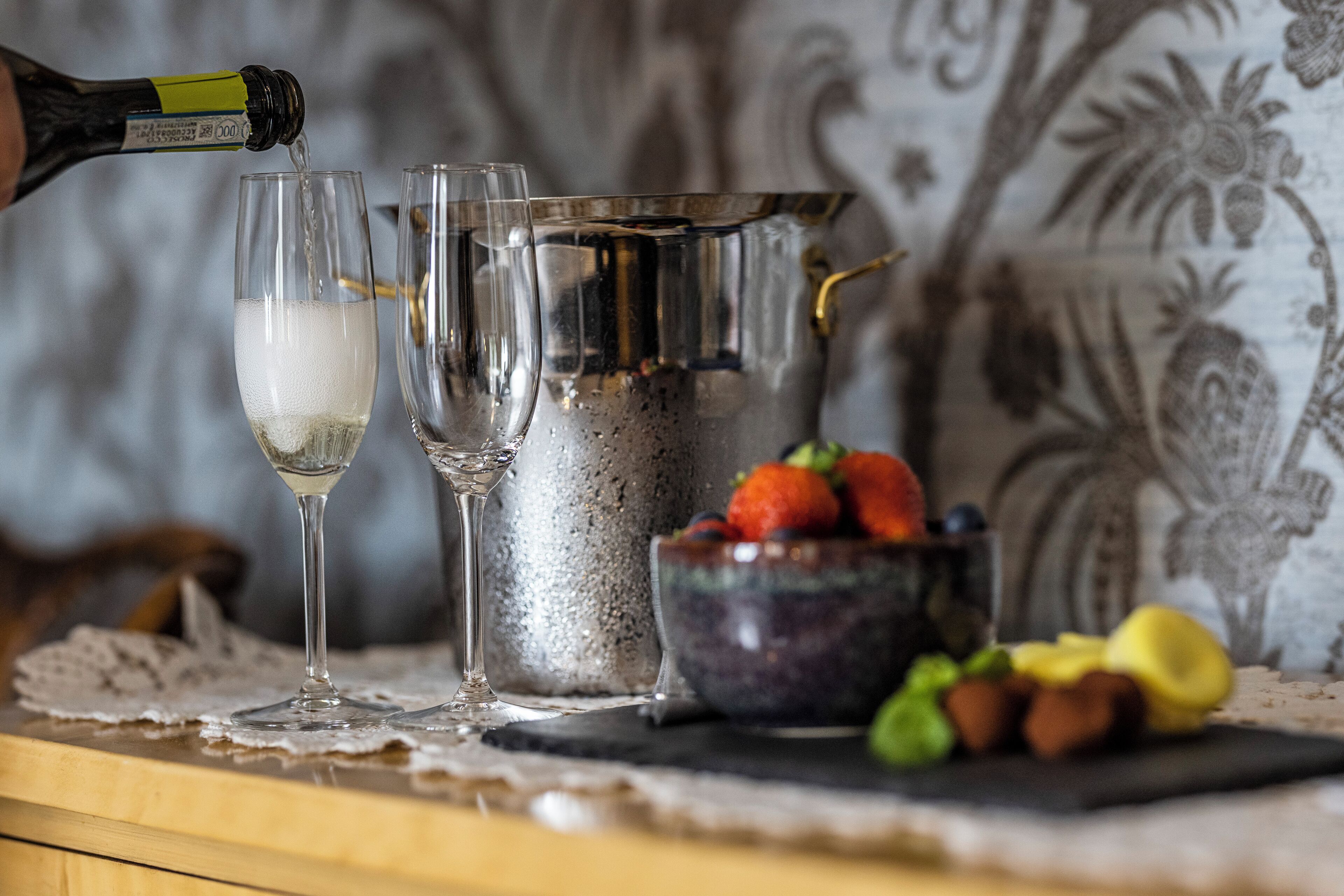 Una bottiglia di champagne che viene versata in due bicchieri, un boccale di metallo e una ciotola di frutta su un tavolo con un sottile centrotavola in pizzo.