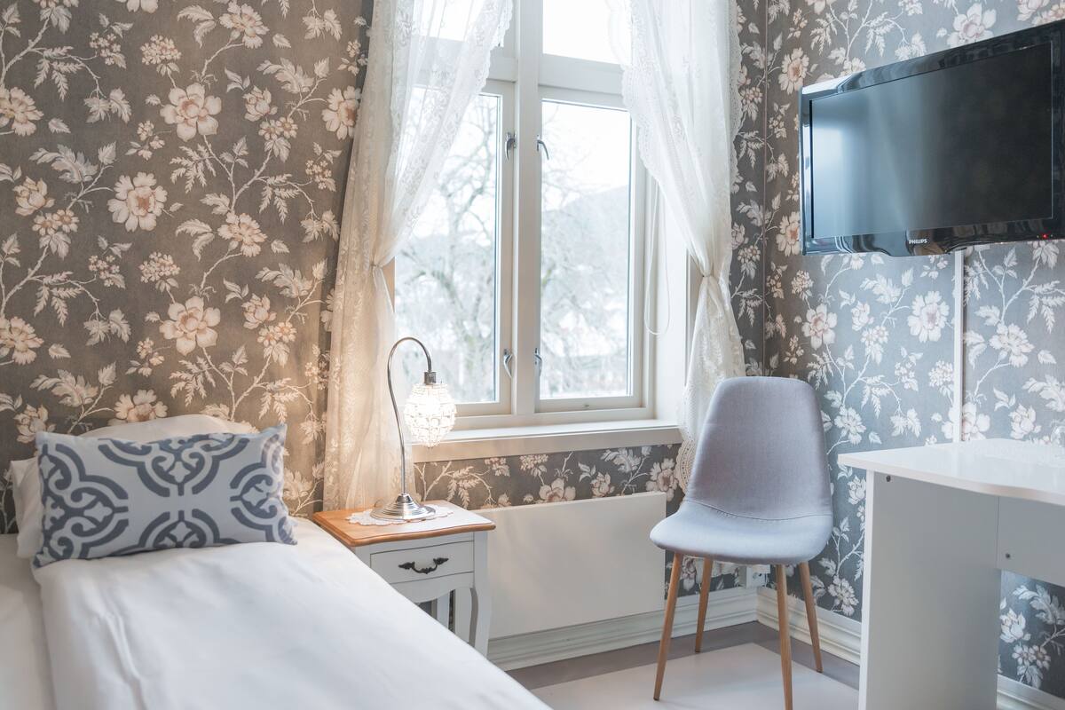 A bedroom with a bed, a desk, a chair, a TV, and a window with curtains.