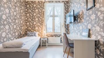 Un dormitorio con cama, escritorio, silla, ventana con cortinas, lámpara y televisor en la pared.
