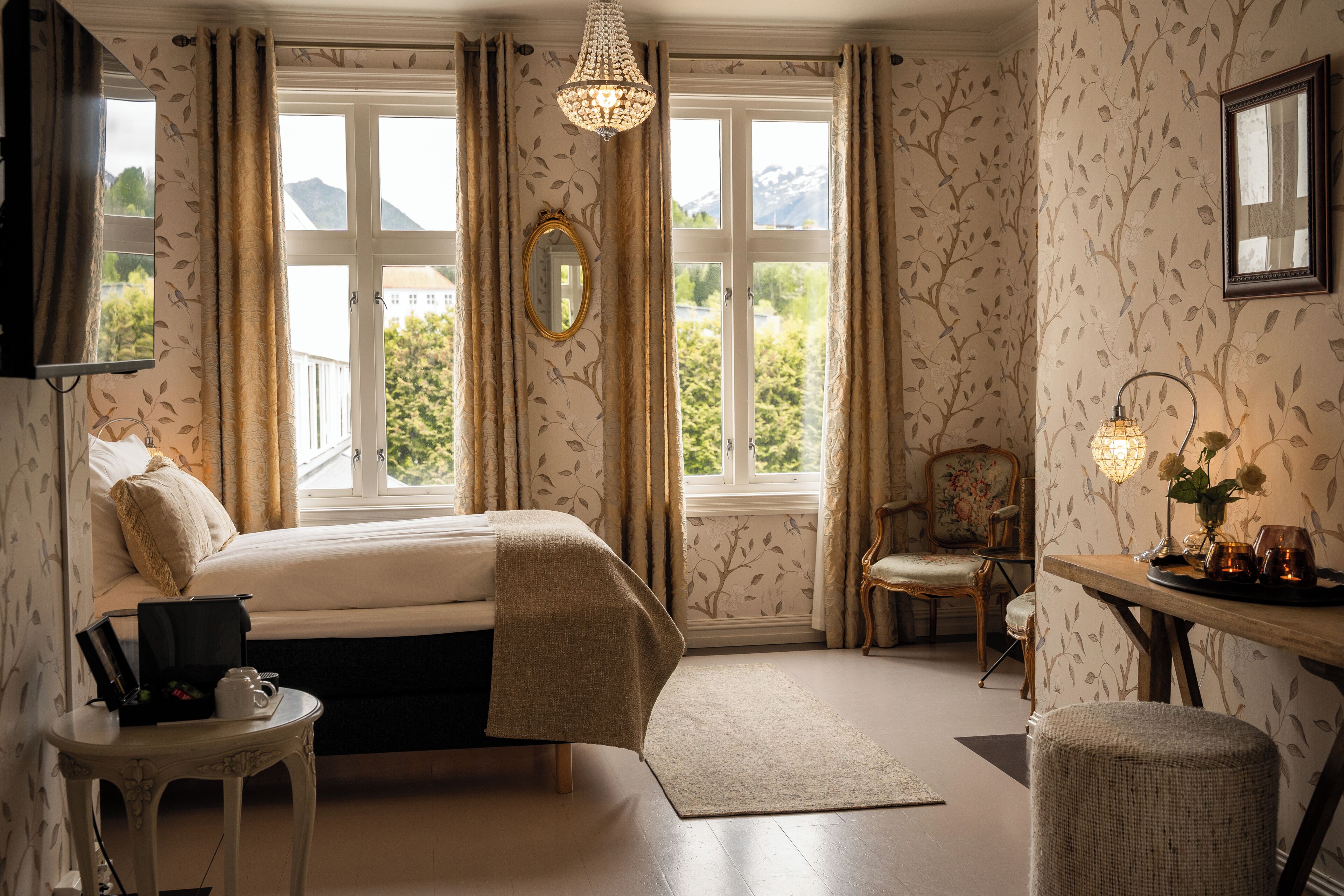 A bedroom with a bed, a bedside table, a chair, a table with a lamp, and a view of the outdoors through large windows.