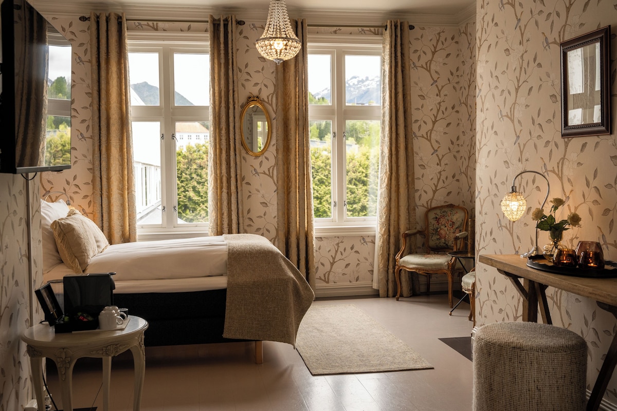 Ein Schlafzimmer mit Bett, Nachttisch, Sessel, Tisch mit Lampe und Blick ins Freie durch große Fenster.