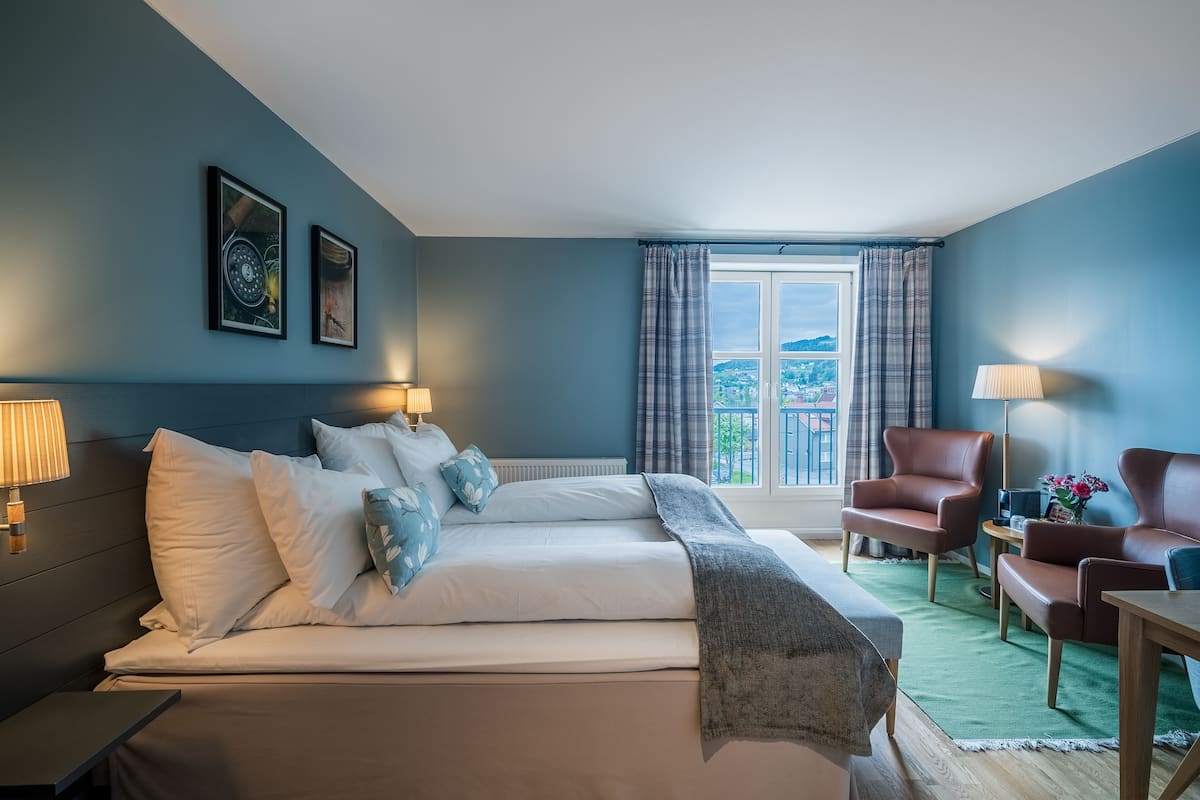A bedroom with a bed, two chairs, a table, a window with curtains, and artwork on the wall.