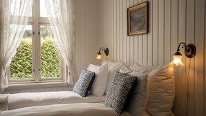 Ein Schlafzimmer mit zwei Betten, weißer Bettwäsche, gemusterten Kissen, einem Fenster mit durchscheinenden Vorhängen und Wandlampen.