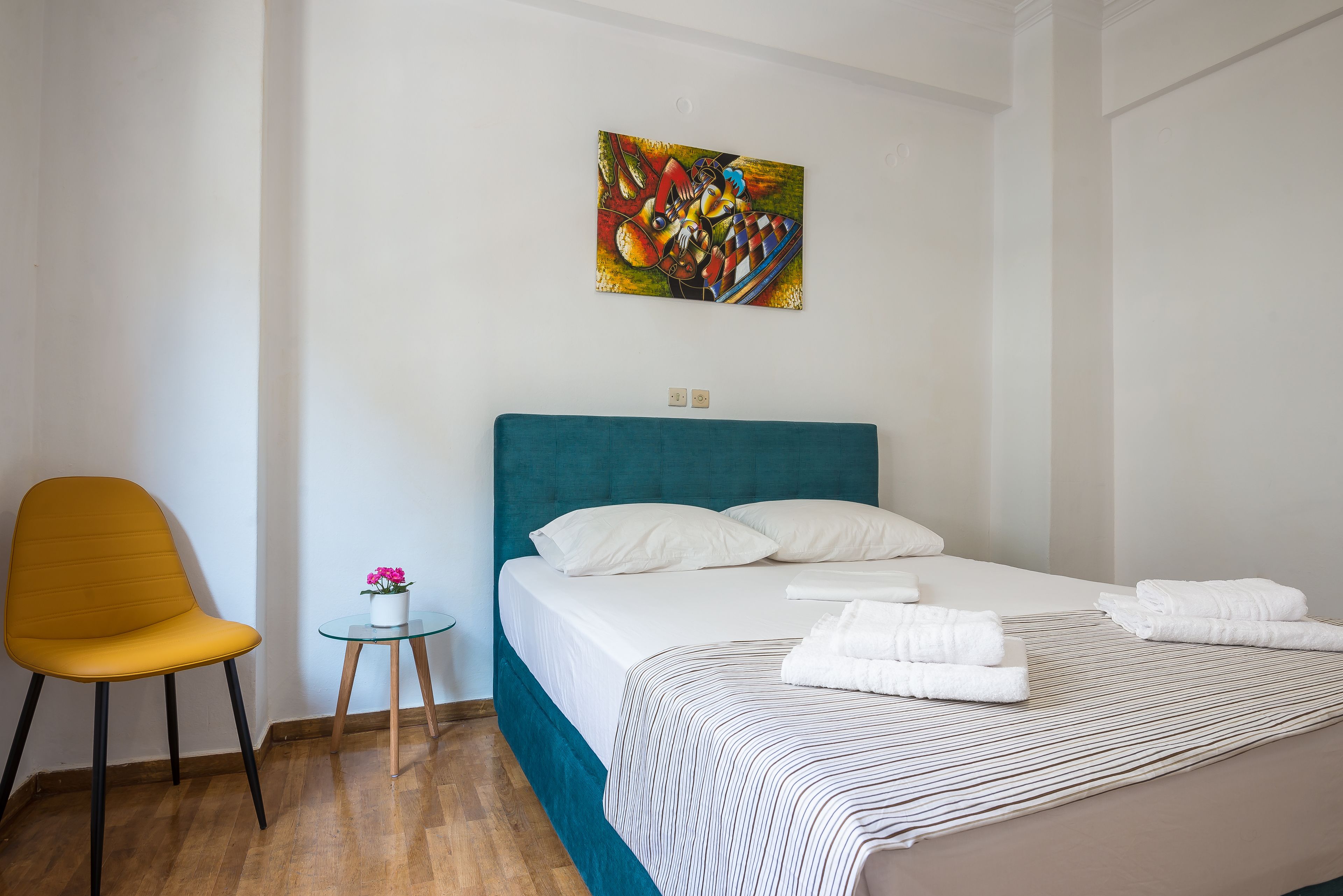 A hotel room with a bed, a chair, a small table with a flower, and a painting on the wall.