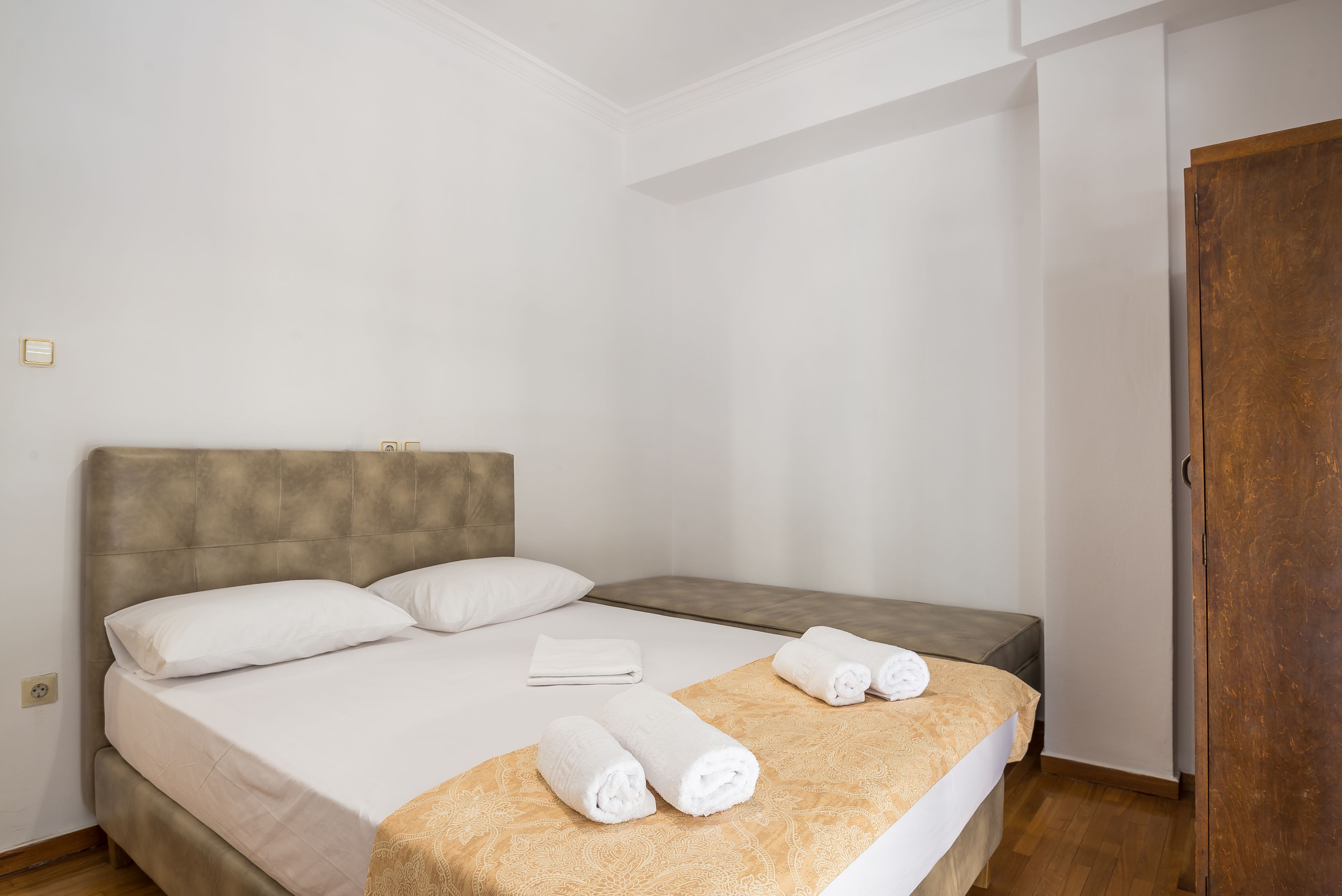 A neatly made bed with white linens and two rolled towels, a wooden headboard, and a wooden door in the background.