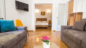 A living room with two sofas, a glass coffee table with flowers, a bed with a headboard, and a TV mounted on the wall. - Victoria Best Apartment (Athens)